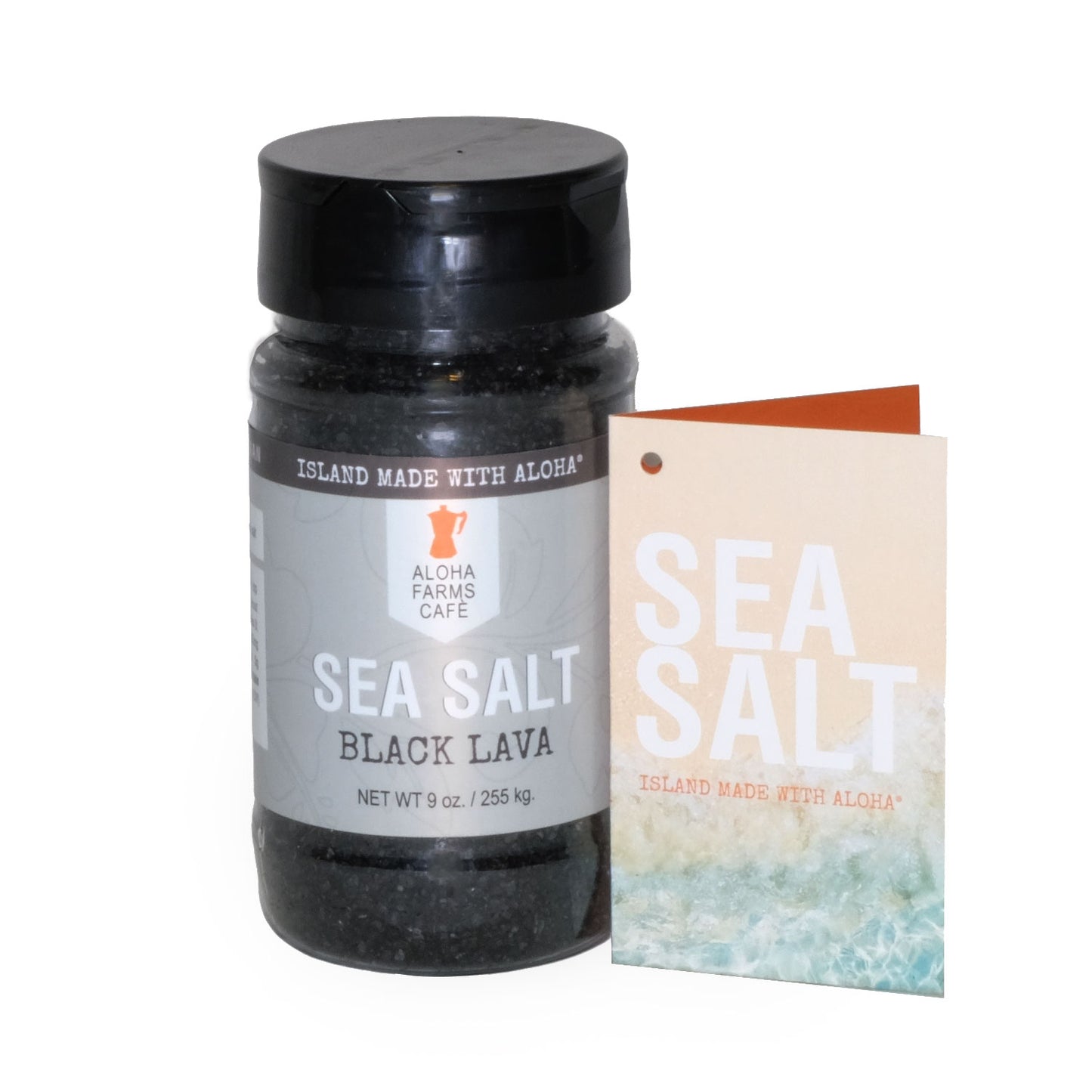A 9 oz. bottle of Aloha Farms Café Volcanic Sea Salt Black Lava, with a label showing the product name and weight, next to a postcard with a beach scene.