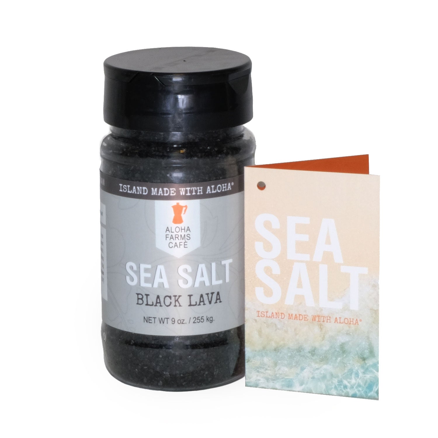 A 9 oz. bottle of Aloha Farms Café Volcanic Sea Salt Black Lava, with a label showing the product name and weight, next to a postcard with a beach scene.