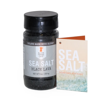 A 9 oz. bottle of Aloha Farms Café Volcanic Sea Salt Black Lava, with a label showing the product name and weight, next to a postcard with a beach scene.